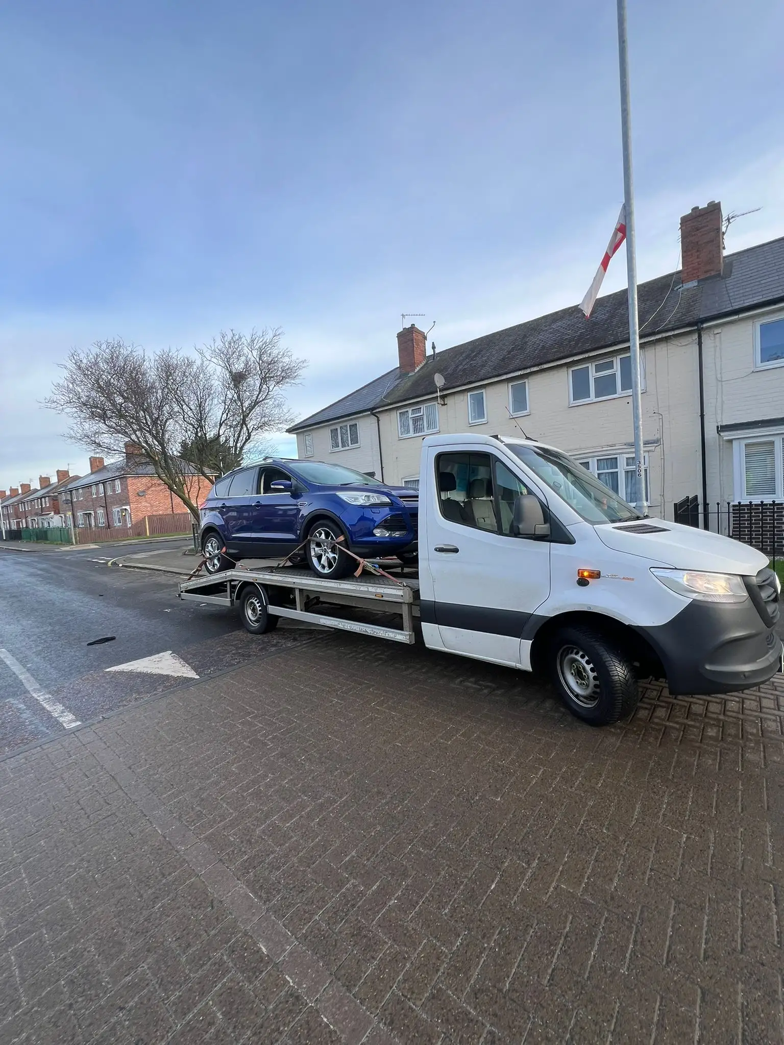 White Sprinter flatbed carrying a blue Ford Kuga on a residential street